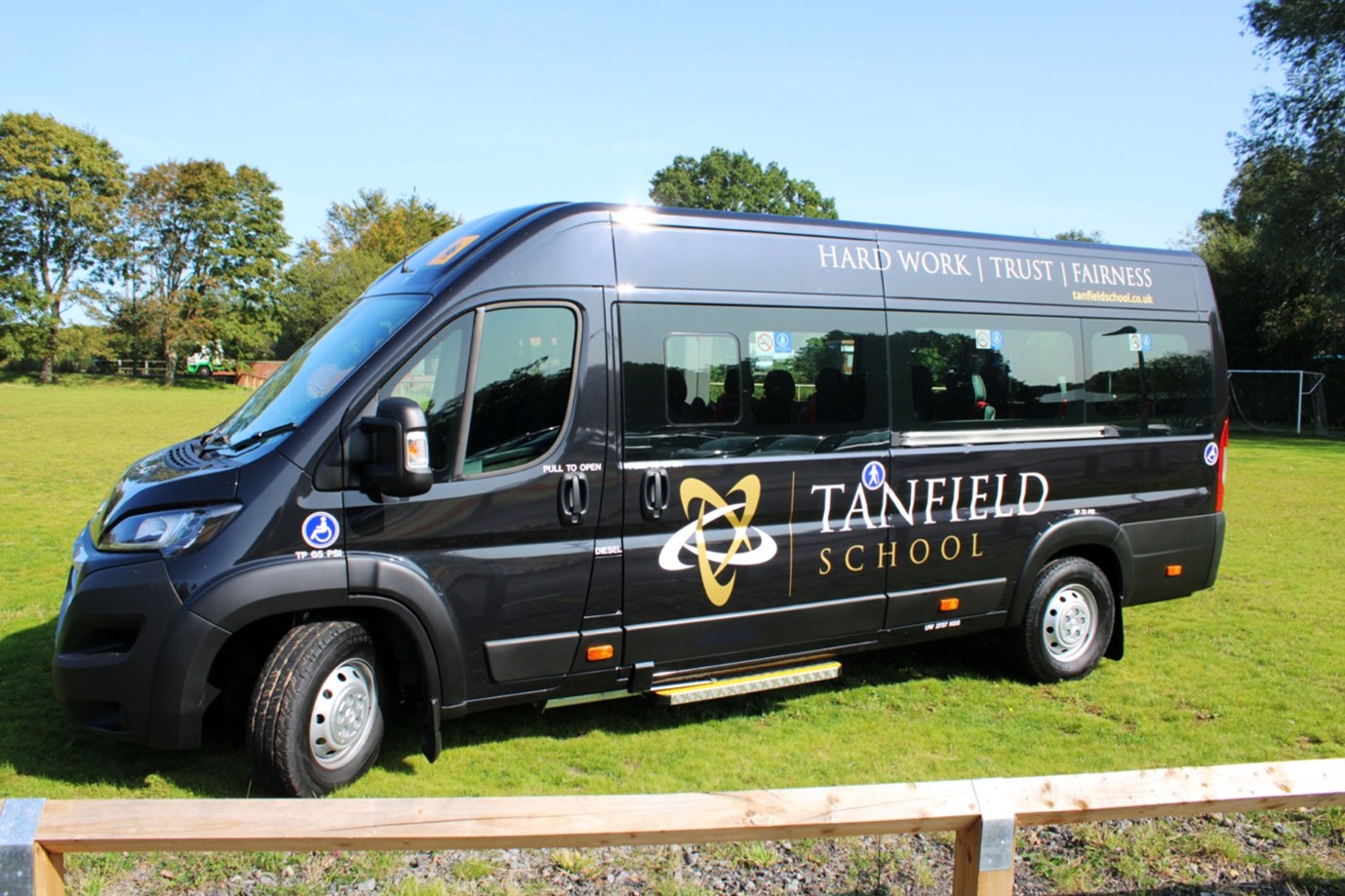 Picture of black minibus on a school field. The mini bus is covered in Tanfield School logos and branding.